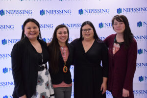 Four women smiling and standing close together in front of a Nipissing University backdrop