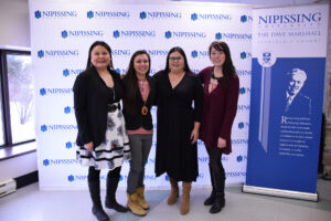 Four women pose together in front of a Nipissing University backdrop beside a Dave Marshall Leadership Awards banner