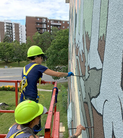 Two painters in hard hats on a lift paint a large mural on the side of a building