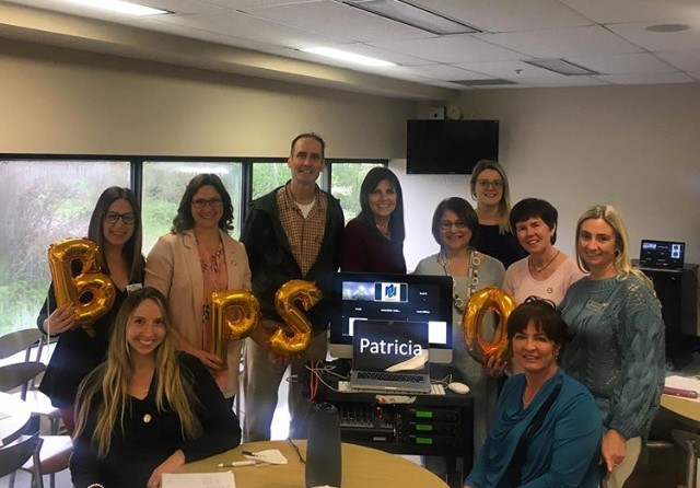 Nine women and one man smiling around a table with gold balloon letters spelling 'BPSO' and a laptop reading 'Patricia'