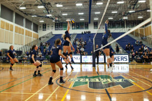 Nipissing women's volleyball players in navy at the net during a game, one jumping to spike the ball