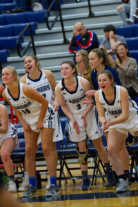 Nipissing women's basketball players in white jerseys cheer and lean forward on the bench