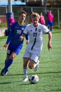 A Nipissing men's soccer player in a white number 23 jersey runs with the ball ahead of a defender in blue