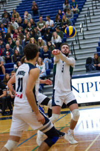 A Nipissing men's volleyball player in white bumps the ball during a match as spectators watch from the stands