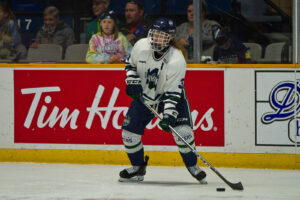 A Nipissing women's hockey player in a white jersey controls the puck at the boards in front of a Tim Hortons ad