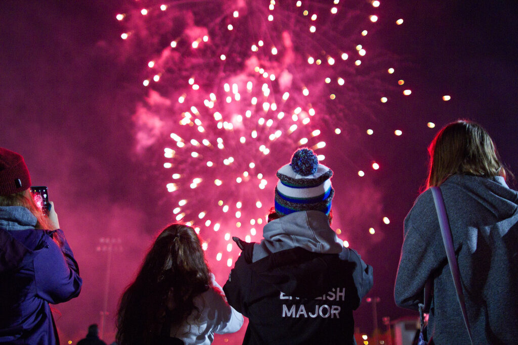 People in jackets and toques watch fireworks in a magenta night sky