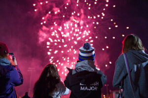 People in jackets and toques watch fireworks in a magenta night sky