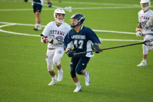 A Nipissing lacrosse player in a navy number 42 jersey runs with the ball while an Ottawa player in white chases him