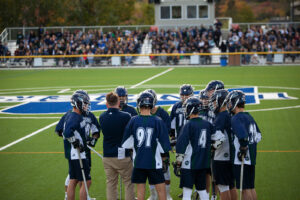 Nipissing lacrosse players in navy huddle with their coach on a turf field before a crowd of spectators