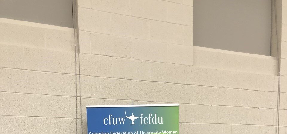 A woman handing an envelope to another woman in front of a Canadian Federation of University Women (CFUW) North Bay banner