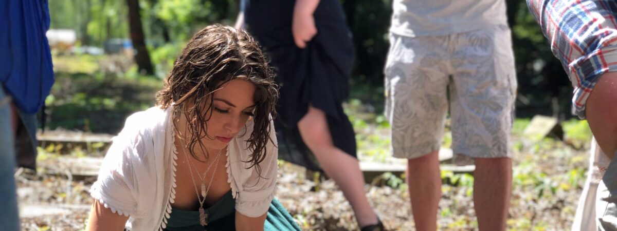 A young woman with wet hair kneeling to touch a flat stone grave marker in a wooded area