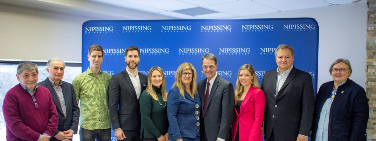 Eleven people in business and casual wear pose in a row in front of a blue Nipissing University backdrop