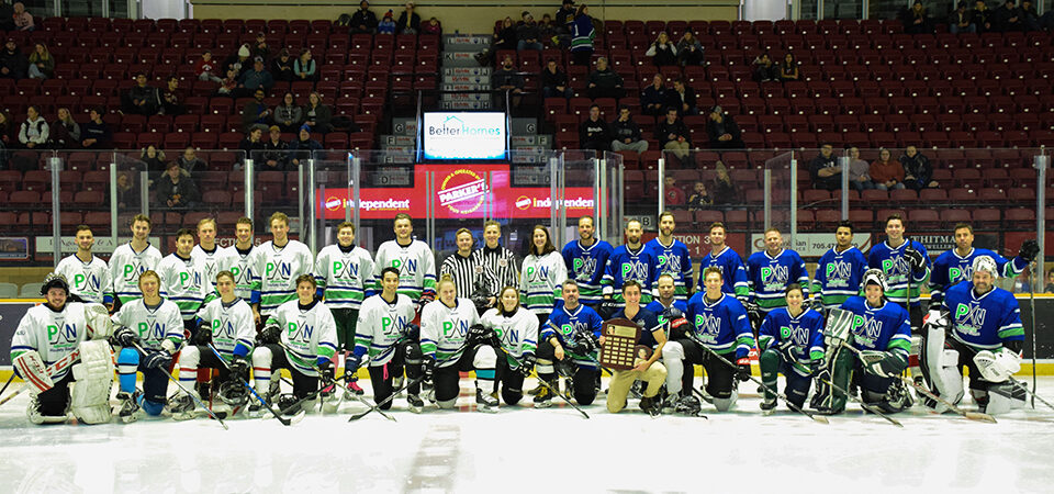 Two hockey teams in white and blue jerseys, along with referees, pose together on the ice in an arena with a trophy in front