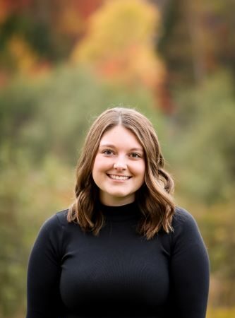 Portrait of a smiling young woman with shoulder-length hair wearing a black sweater, against a blurred autumn background