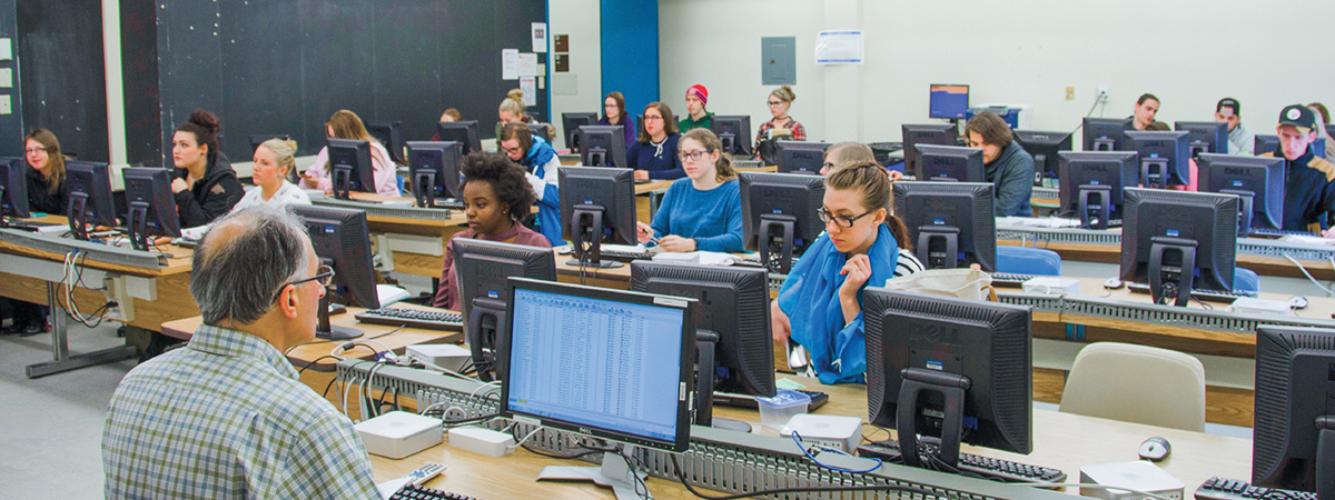 Students at rows of desktop computers in a computer lab, with an instructor in the foreground
