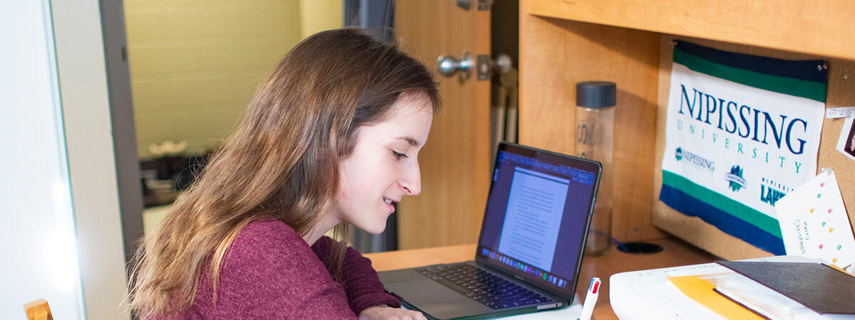 A young woman writing at a desk next to an open laptop, a printer and a Nipissing University pennant