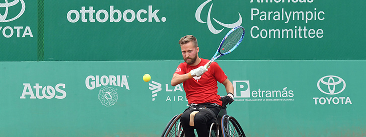 A wheelchair tennis player in a red shirt returns a shot on a clay court in front of sponsor banners