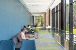Students sitting and working in blue and green lounge seating along a bright university hallway with a glass wall