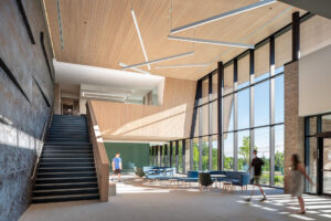 Bright atrium with a wide staircase, wood-panelled ceiling and tall windows, with students walking through and sitting on blue sofas