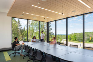 Students working around a long conference table in a corner room with floor-to-ceiling windows overlooking trees and a road