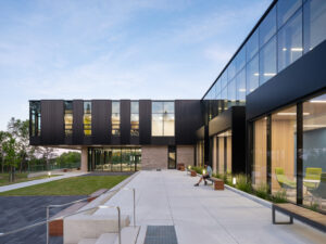 Modern two-storey building with black panel cladding and glass walls overlooking a paved courtyard and lawn