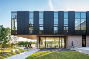 Modern two-storey building with black panel cladding, brick base and large windows, seen from a grassy lawn