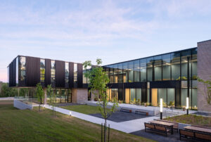 Modern building with a courtyard, benches and young trees at dusk, its glass walls lit from inside
