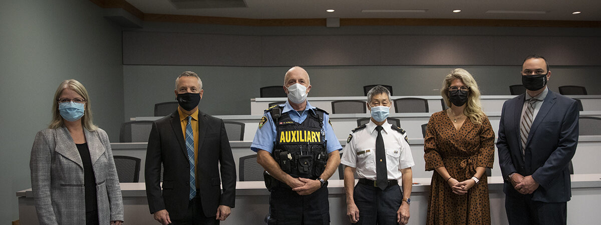 Six masked people, including two uniformed police officers, stand in a row in a lecture room