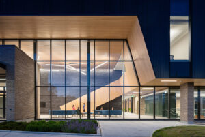 Glass entrance of a modern building lit at dusk, showing a wooden staircase and blue lounge seating inside
