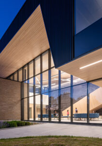 Close-up of a modern building's angled wood-panelled overhang and glass wall, lit at dusk with people seated inside