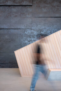 A blurred person walking past a wooden angled wall panel set against a dark textured wall