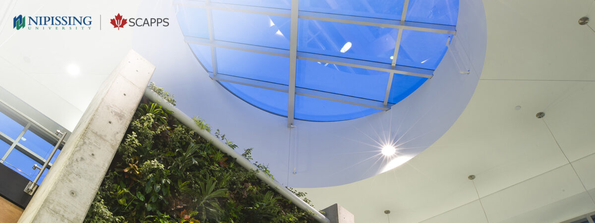 Looking up at a living green wall beside a concrete column under a skylight in a university atrium, with Nipissing University and SCAPPS logos