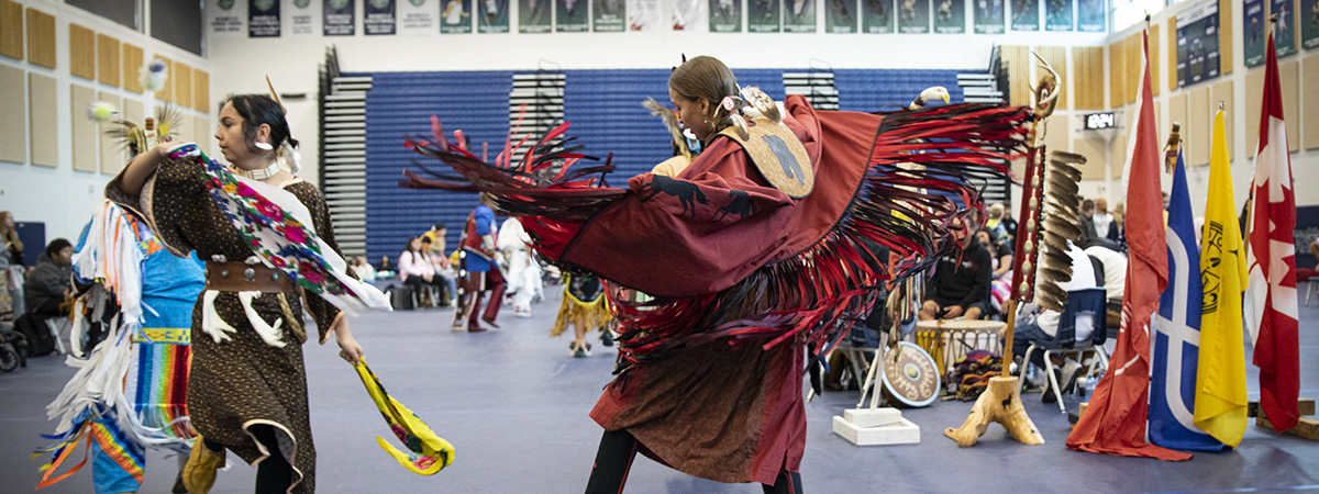 Dancers in regalia performing at a powwow in the university gym, with flags standing at the side