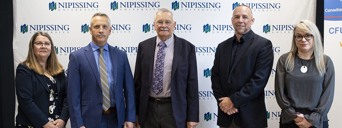 Five people in business attire stand in a row in front of a Nipissing University backdrop
