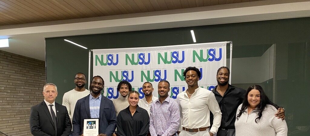 A group of ten men and women pose in front of a green and blue NUSU backdrop, one man holding a framed plaque