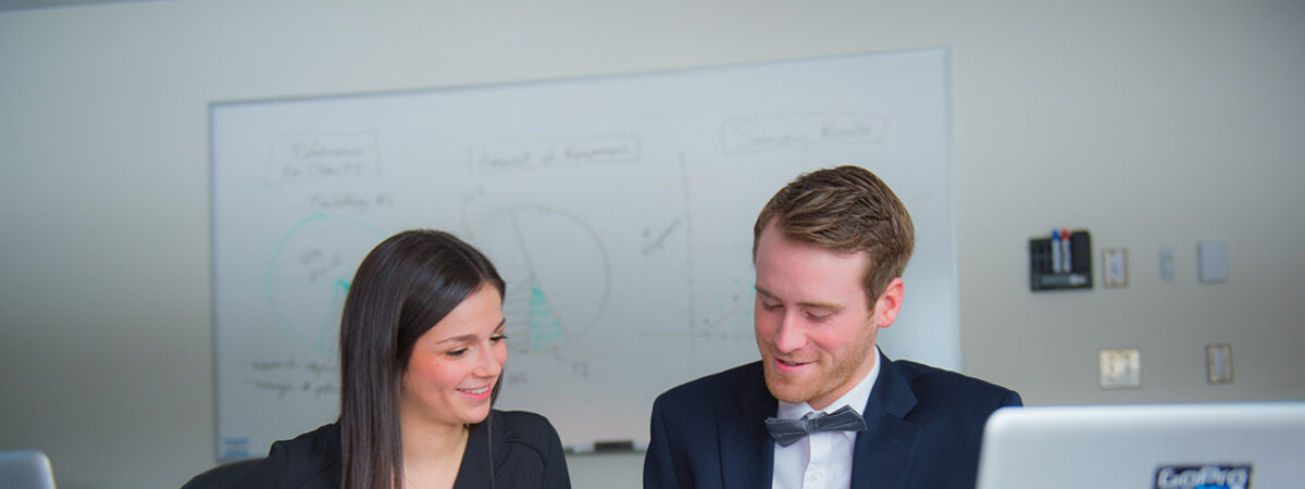 A woman and a man in a bow tie working together at a table with laptops and papers, with equations on a whiteboard behind them