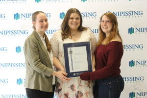 Three smiling women in front of a Nipissing University backdrop holding a framed Dave Marshall Leadership Award certificate together