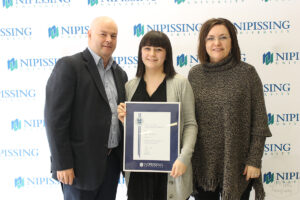 A young woman holding a framed Dave Marshall Leadership Award certificate stands between a man in a navy blazer and a woman in a grey poncho