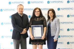 A young woman in a black lace dress holding a framed Dave Marshall Leadership Award certificate stands between a man in a navy blazer and a woman in a tweed jacket