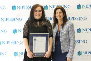 A young woman holding a framed Dave Marshall Leadership Award certificate stands beside a smiling woman in a tweed jacket
