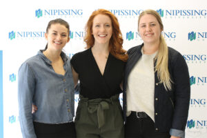 Three smiling women with their arms around each other in front of a Nipissing University backdrop
