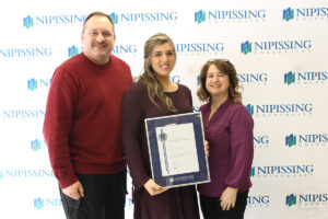 A woman holding a framed Dave Marshall Leadership Award certificate stands between a man in a red sweater and a woman in a purple blouse in front of a Nipissing University backdrop