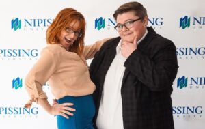 Two people pose playfully in front of a Nipissing University backdrop; one has red hair and glasses, the other wears a blazer and rests a hand on their chin