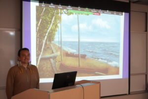 A man in a tan sweater standing at a podium in front of a projection screen showing a vintage lakeside beach scene