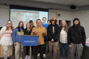 A group of students and instructors posing for a group photo beside a Nipissing University podium in a classroom
