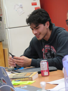 A smiling young man in a hockey hoodie sits at a table using a craft knife, with a drink can and books in front of him