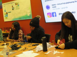 Three students crafting at a table covered in felt scraps and supplies in front of a screen displaying social media information for an Indigenous student group