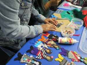 Close-up of hands assembling felt dolls decorated with buttons, with glue and a bowl of beads on a blue table