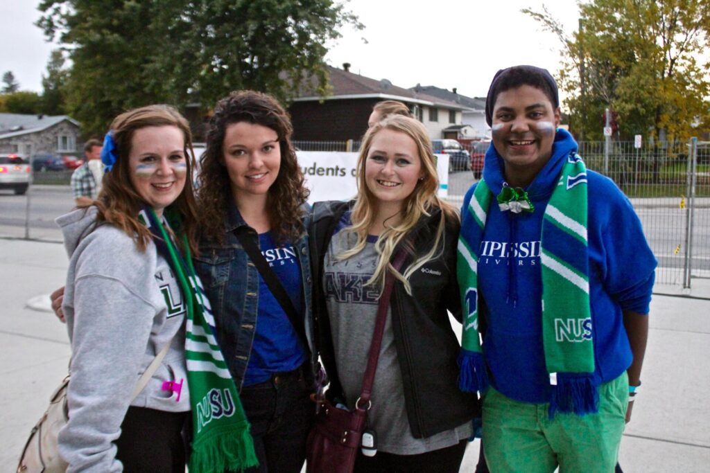 Four smiling people wearing Lakers and NUSU scarves and hoodies pose together outdoors