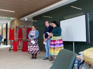 Two women in regalia and a man stand in front of a projection screen in a hall, beside a rack of red dresses